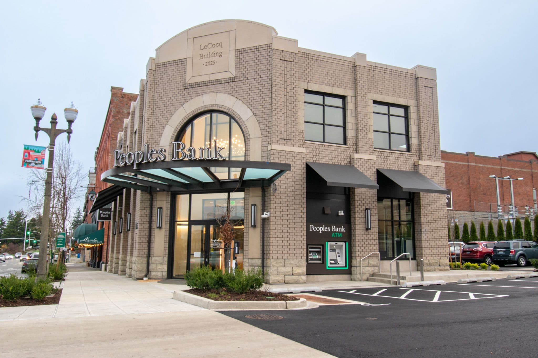Exterior view of a contemporary Peoples Bank branch in a brick commercial building, featuring an arched entryway, glass canopy, ATM access, and landscaped sidewalk in a vibrant downtown setting. The photo highlights modern architecture, pedestrian‑friendly design, and convenient customer access.